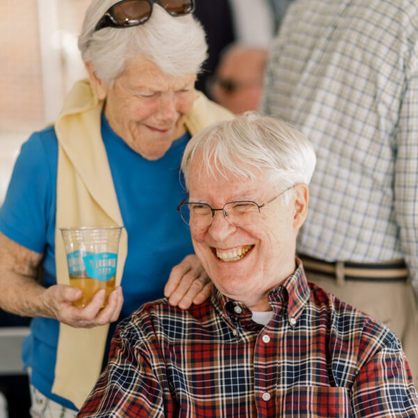 Two elderly men at a casual gathering; one seated in a red plaid shirt smiling, the other standing behind with sunglasses on his head and a beer in hand