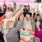 Elderly group raising their hands in celebration in a circle at an outdoor concert under a tent, with a band playing on stage behind them.