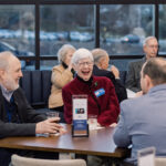 Group of smiling senior adults seated around a table at a social event, chatting and drinking beverages in a bright lounge.