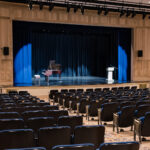 Empty theater with blue stage curtains, a grand piano and a lectern on stage, rows of black seats in the foreground.