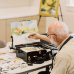 Older man with white hair and glasses paints a watercolor landscape at a tabletop easel in a bright studio session.