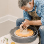 Older man in a blue plaid shirt and apron shapes clay on a pottery wheel, concentrating on the piece in progress.