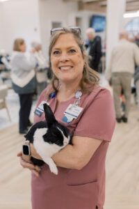 Smiling woman in pink scrubs cradles a black-and-white cat in a busy veterinary or shelter lobby.