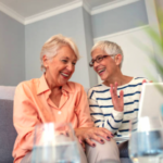 Two elderly women sit on a sofa, smiling and chatting in a cozy living room setting.