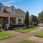 Neighborhood scene: people gardening in front of a brick house with white columns and hanging flower baskets on a sunny day