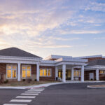 Front view of a brick-and-white community center with columns and large windows at sunset, with a striped crosswalk in the foreground.