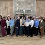 Group of about 20 adults posing for a photo in a formal room with a stone fireplace and wood-paneled walls; several wear name badges and smart clothing.