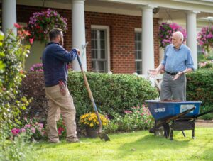 Two men gardening outside a brick house, one holding a long tool near a wheelbarrow with blooming flowers nearby.