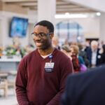 Smiling Black man with glasses in a maroon sweater at a crowded indoor event, wearing a name badge that says 'New Employee'