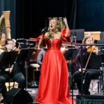 Female singer in a red gown performs with an orchestra on stage, microphone in hand.
