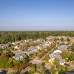 Aerial view of a suburban neighborhood with rows of houses, tree-lined streets, and a distant tree line under a clear blue sky.